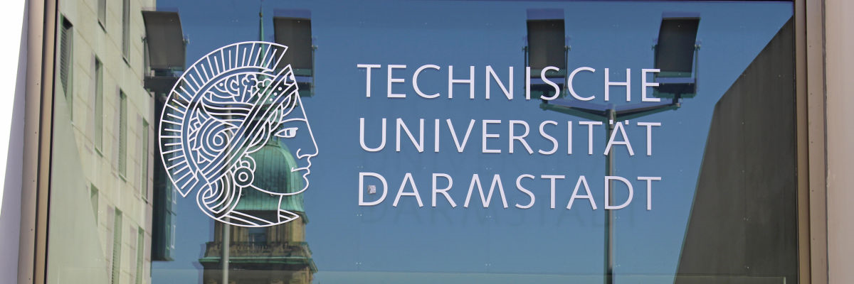 Das Athene-Logo der TU Darmstadt an einer Glasfront, in der sich Laternen und andere Gebäude auf dem Karolinenplatz spiegeln